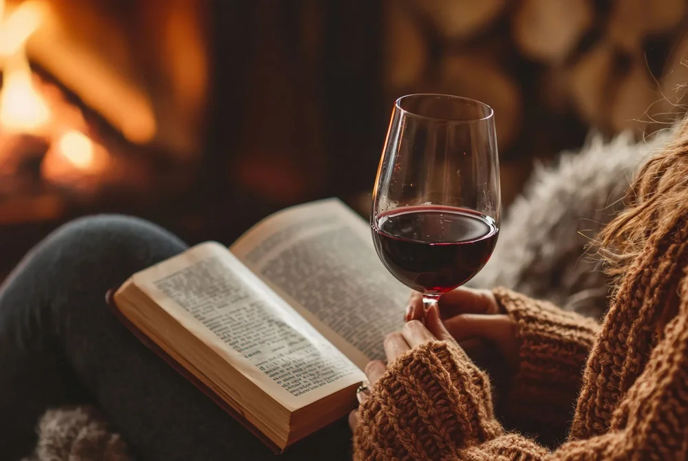 An open book and a glass of red wine by the fireplace, from Decant